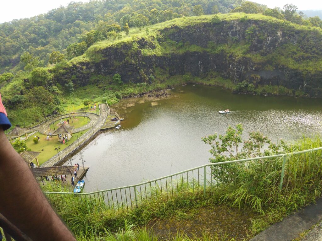 A quiet lakeside view near Idukki, with visitors walking along stone paths and small gazebos by the water, while a couple enjoys boating under the shade of green cliffs.