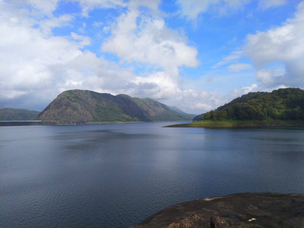 A serene view of Idukki reservoir surrounded by green hills and rocky slopes under a blue sky with drifting clouds, reflecting calm waters below.