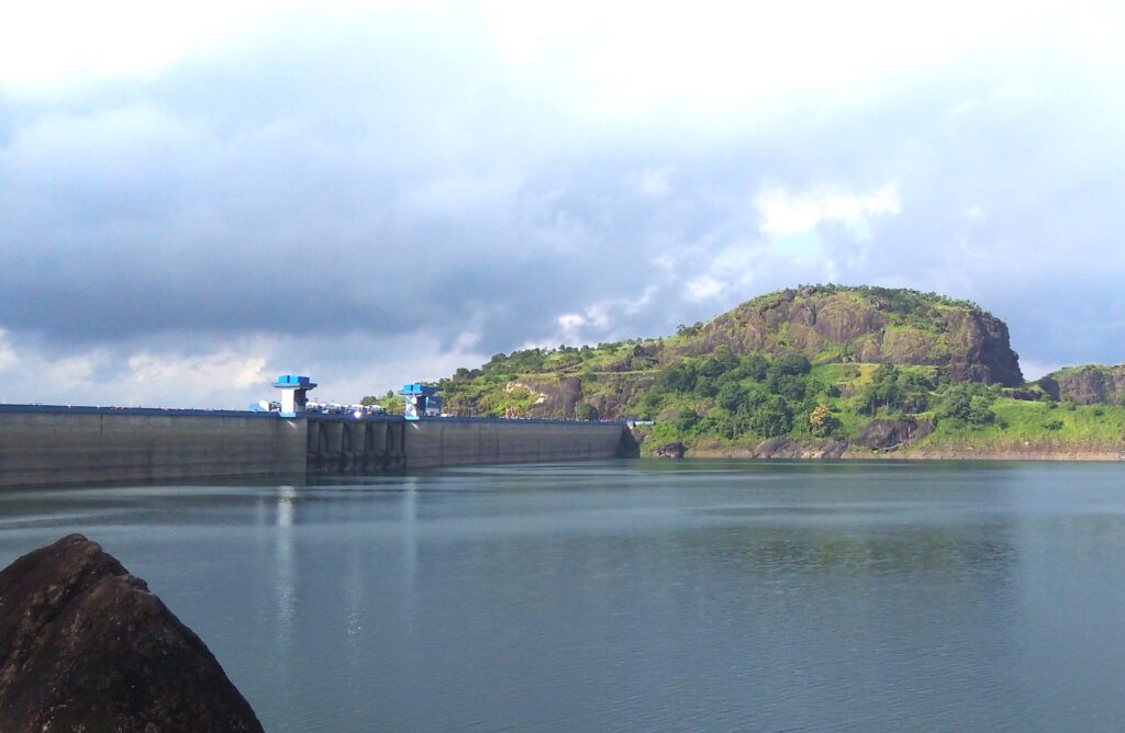View of the Idukki Dam and reservoir surrounded by rocky green hills under a cloudy sky, with calm water reflecting the landscape.