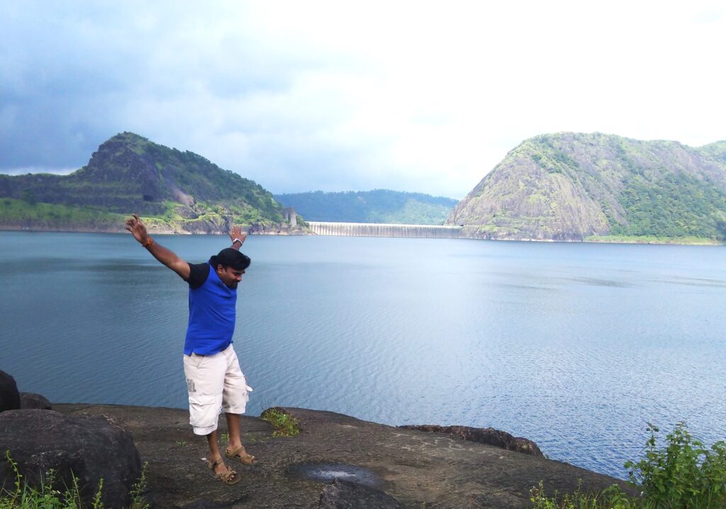 A man in a blue shirt and white shorts joyfully jumping on a rocky edge overlooking the Idukki Dam and its vast reservoir, surrounded by green hills under a cloudy sky.