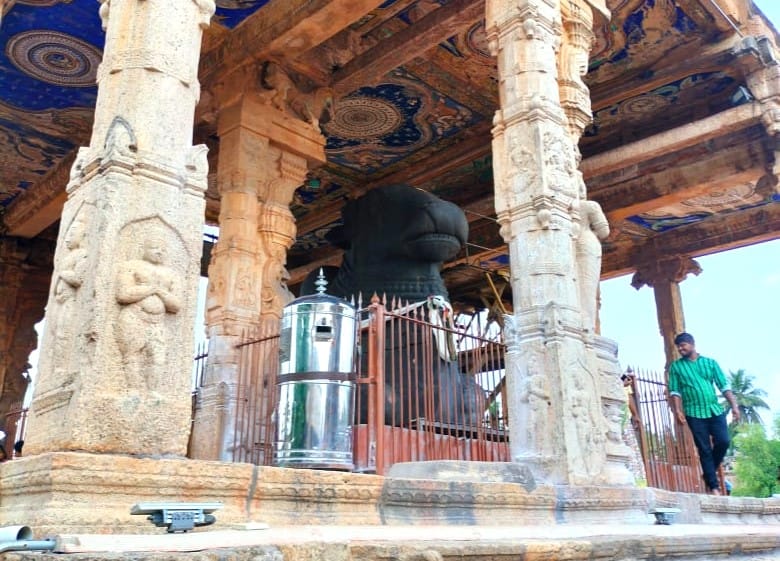 Stone pavilion with intricately carved pillars and a vibrantly painted ceiling, housing a large black Nandi statue enclosed by a metal fence in Thanjavur ; a man in a green shirt walks past on the right side of the scene.