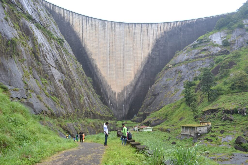 Visitors walking along a narrow path at the base of the towering Idukki Arch Dam, surrounded by steep rocky cliffs covered in green moss and grass after rain.