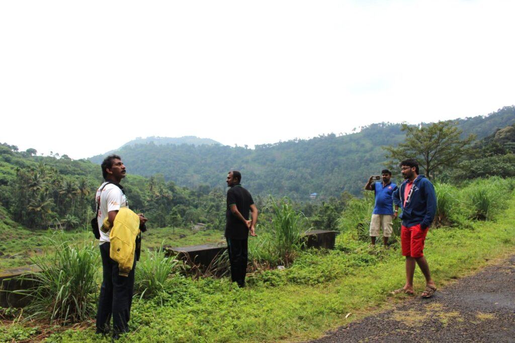 A group of travelers standing on a hillside road surrounded by lush greenery, looking out over the misty valleys of Idukki after rain.
