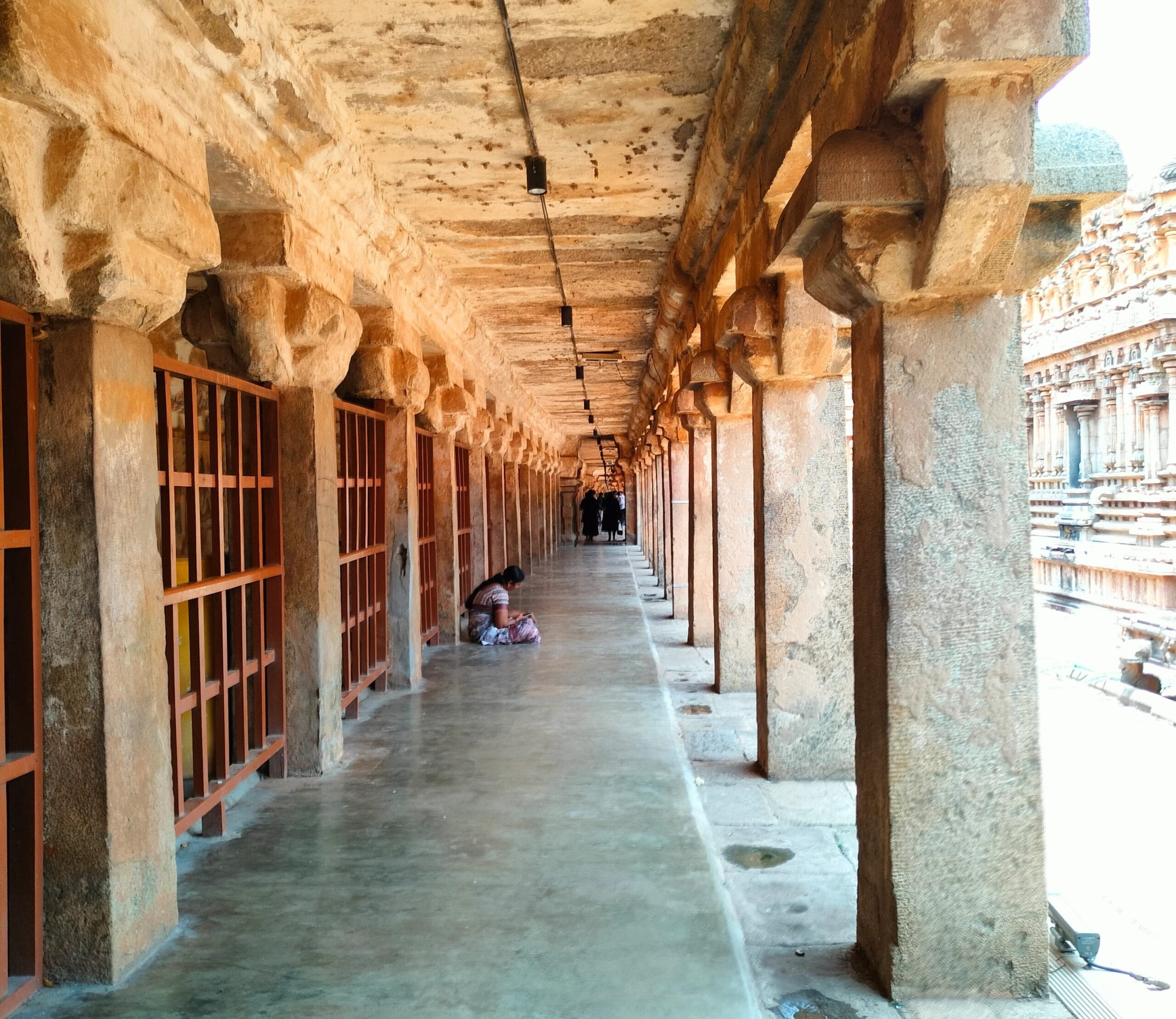 Long stone corridor inside the Brihadeeswarar Temple at Thanjavur, lined with thick carved pillars and a smooth walkway, with a woman seated on the floor and a few visitors visible in the distance.