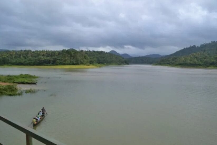 Wooden boat with people crossing a wide river surrounded by green hills during monsoon.
