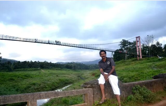 Man sitting near a suspension hanging bridge across a green valley, used by villagers as a lifeline during monsoon in rural Kerala.