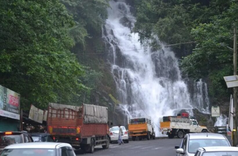 Large waterfall flowing beside a mountain road with trucks and vehicles passing during monsoon.