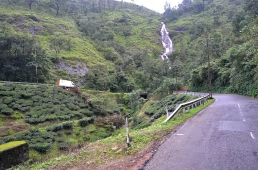 Waterfall flowing down a green hill near a winding road and tea plantations during monsoon.