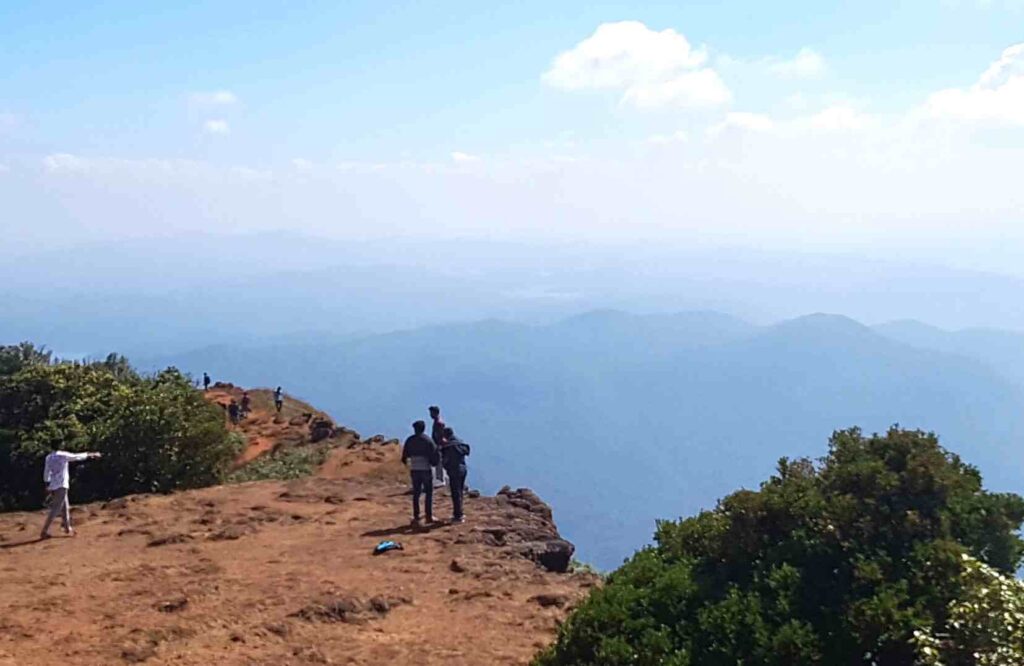 Visitors standing at the cliff edge near Sarvanjapeetha overlooking layered mountain ranges