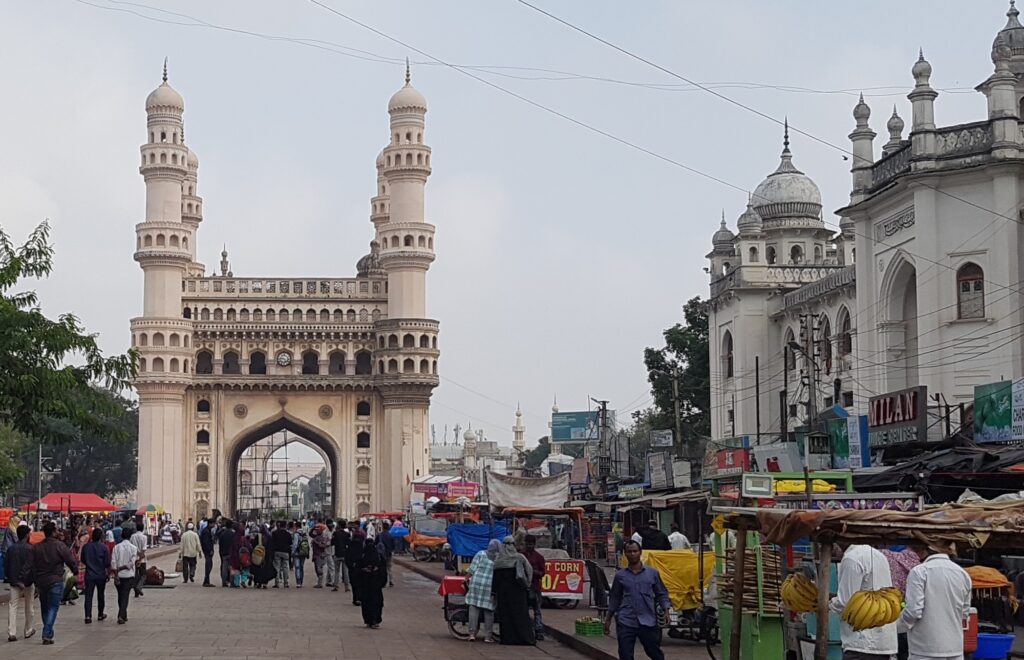 Front view of Charminar in Hyderabad with busy market streets, people, and vendors in the foreground.