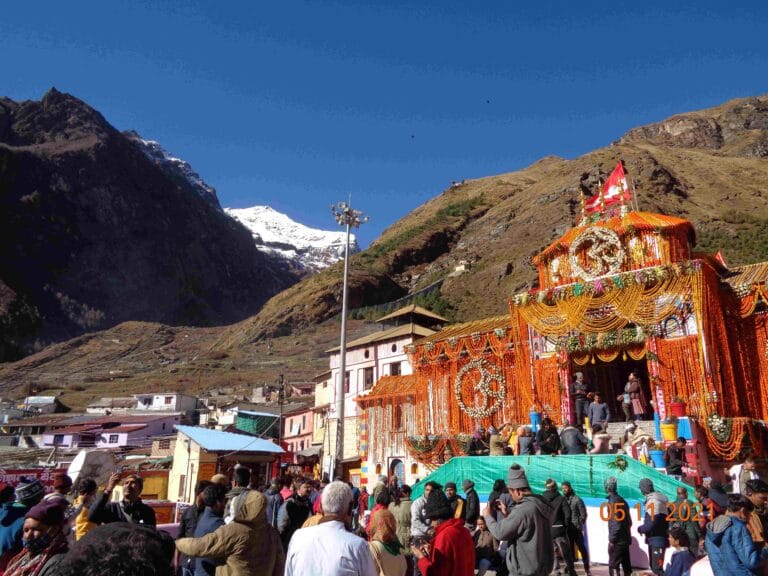 Crowd of pilgrims gathered in front of the brightly decorated Badrinath Temple, surrounded by tall brown mountains and a snow-covered peak under a clear blue sky.