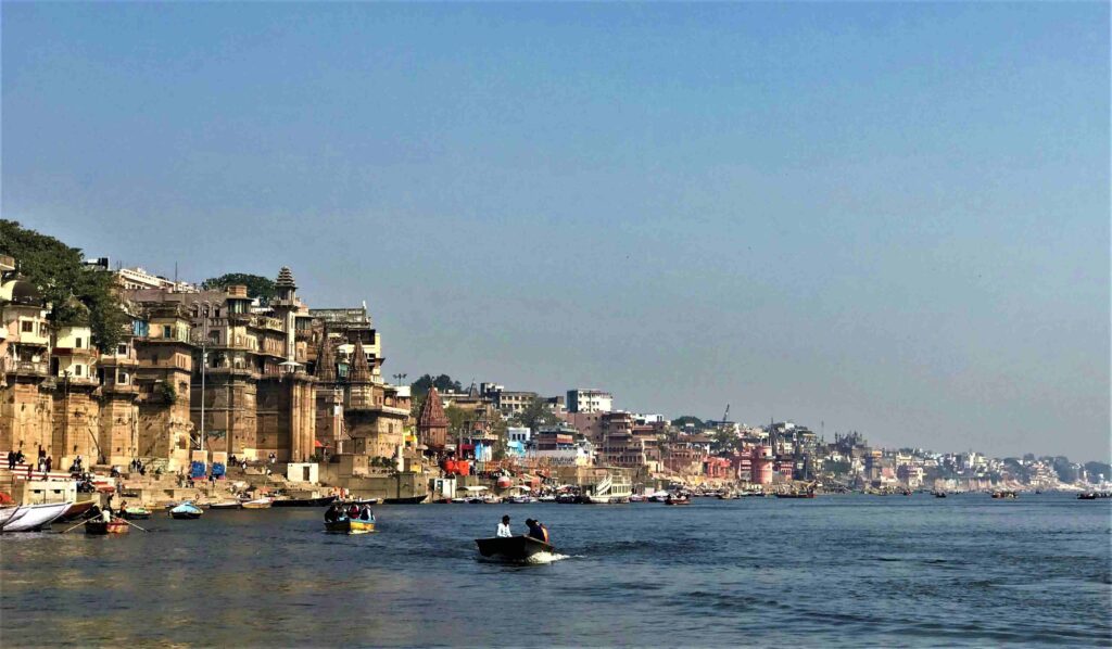 Panoramic view of the Varanasi ghats along the Ganga River, with boats on the water and historic riverside buildings under a clear blue sky.