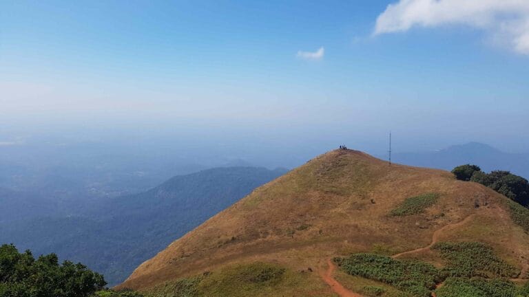 A wide view of Kudajadri hilltop with trekkers standing on the peak under a clear blue sky.