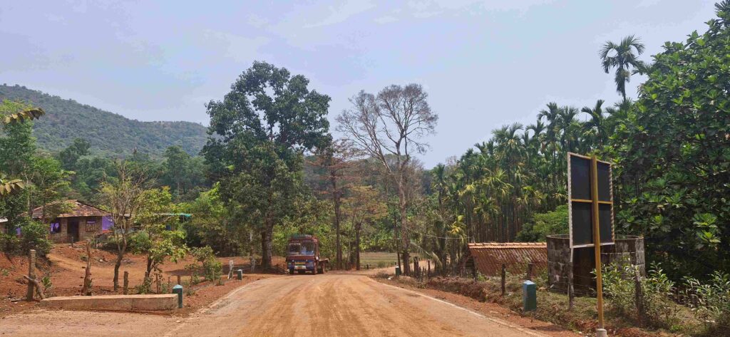 A rural mud road leading towards the Kudajadri checkpost, with hills in the background, village houses on one side, and tall betel nut trees lining the path.