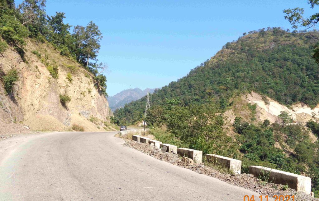 A winding mountain road with steep rocky cliffs on one side and green hills on the other, captured on a bright clear day in the Uttarakhand Himalayas.