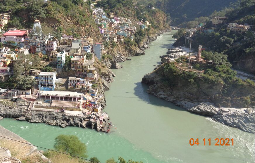 A wide view of the Ganga River at Rishikesh with buildings perched on the hillside, clear green water, and rocky riverbanks under bright sunlight.