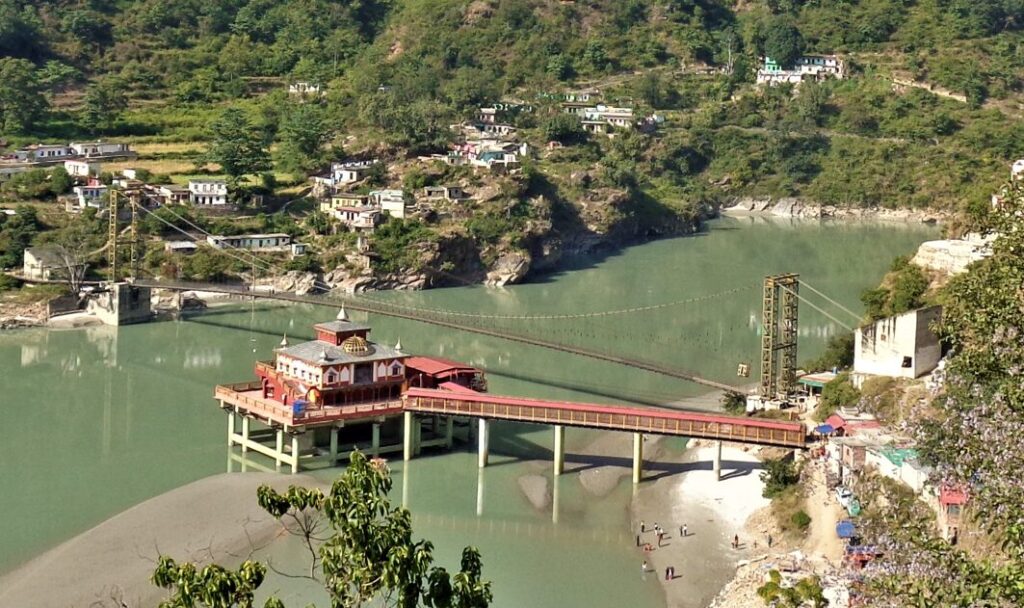 Dhari Devi Temple built on pillars in the middle of the Alaknanda River near Srinagar, Uttarakhand, with surrounding hills and suspended footbridge.”