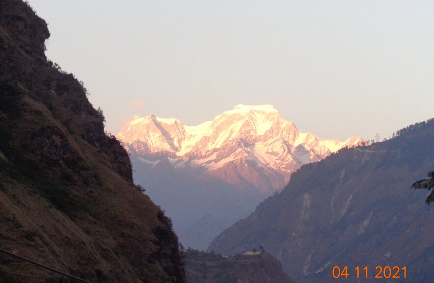 Snow-covered Himalayan peaks glowing in the orange evening light near Badrinath, with dark foreground mountains framing the scene.