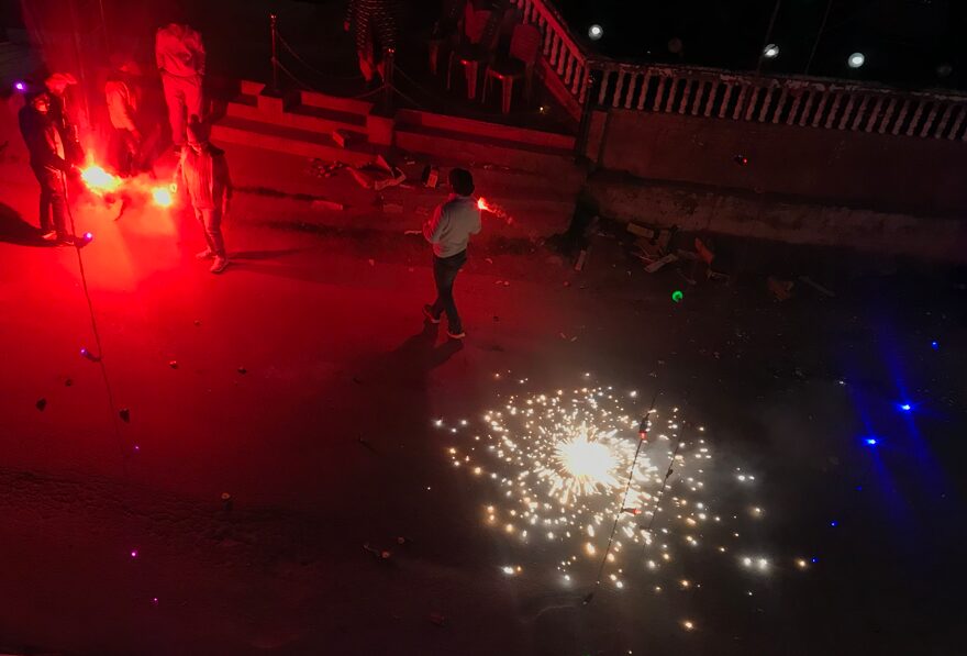 “People celebrating Diwali at night in Joshimath, holding bright red flares while a ground firework sparkles on the street.”