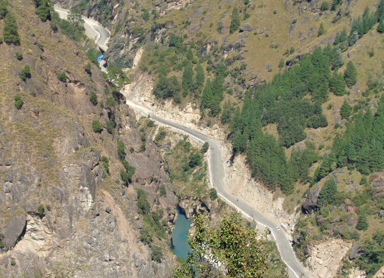 A winding mountain road running through a deep rocky valley with steep cliffs, green patches of pine trees, and a turquoise river flowing below in a remote Himalayan landscape.