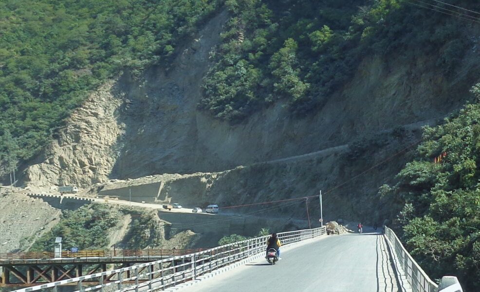 Winding mountain road built along a steep Himalayan cliff with vehicles and a scooter traveling through rocky terrain on the Badrinath route.
