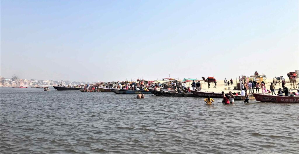 Opposite bank of the Ganga in Varanasi, with people bathing, boats lined along the shore, and the city’s ghats visible across the river.