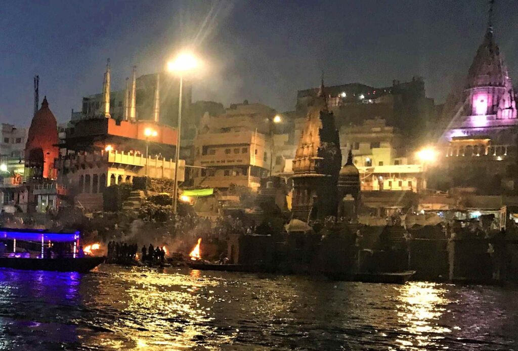Manikarnika Ghat in Varanasi with temple spires, stacked funeral firewood, smoke from cremation rituals, crowded steps, and boats along the Ganga