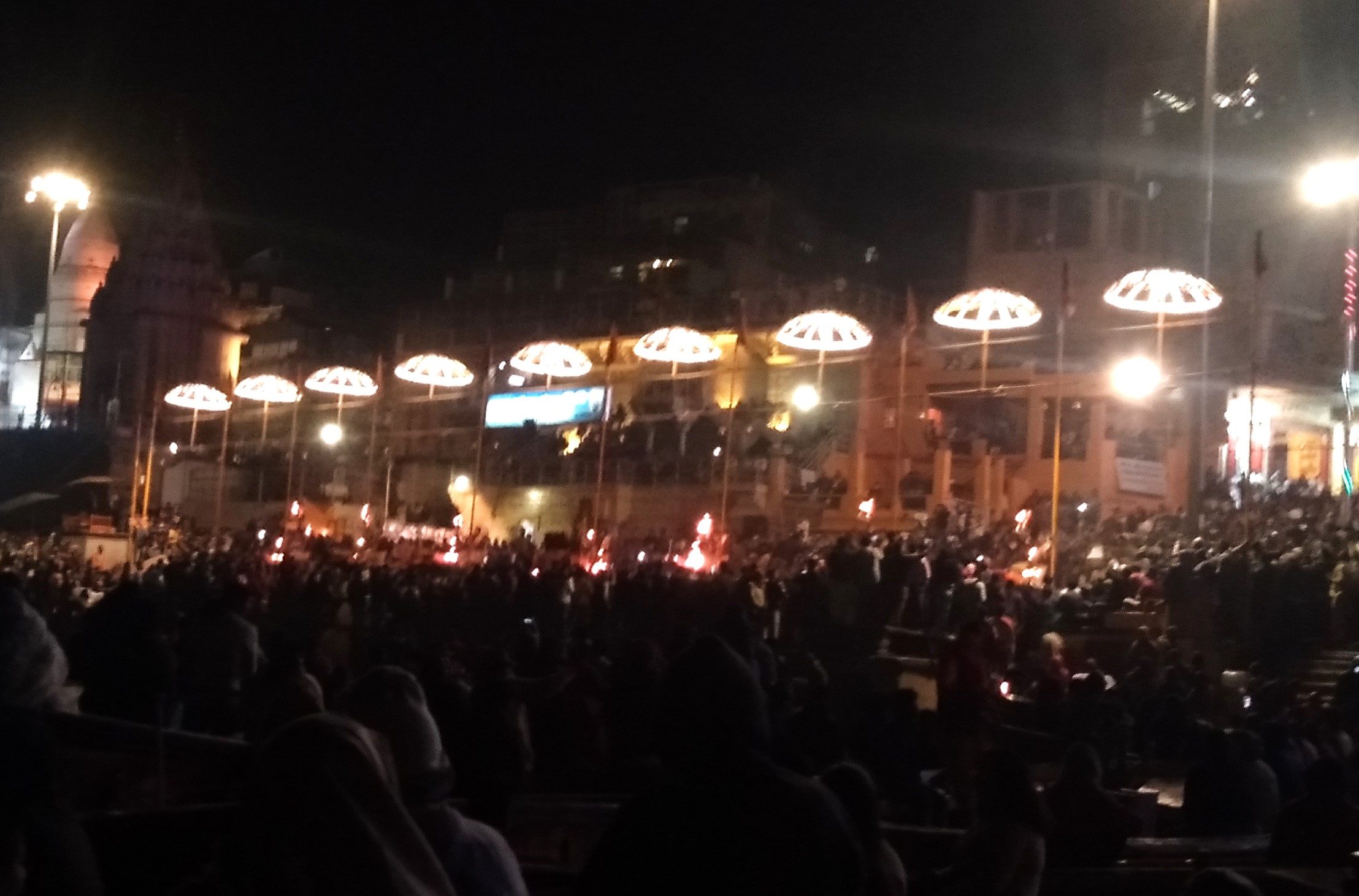 Crowds gathered at Varanasi’s ghat during nighttime Ganga Aarti, with priests performing rituals holding flaming lamps under bright illuminated canopies.
