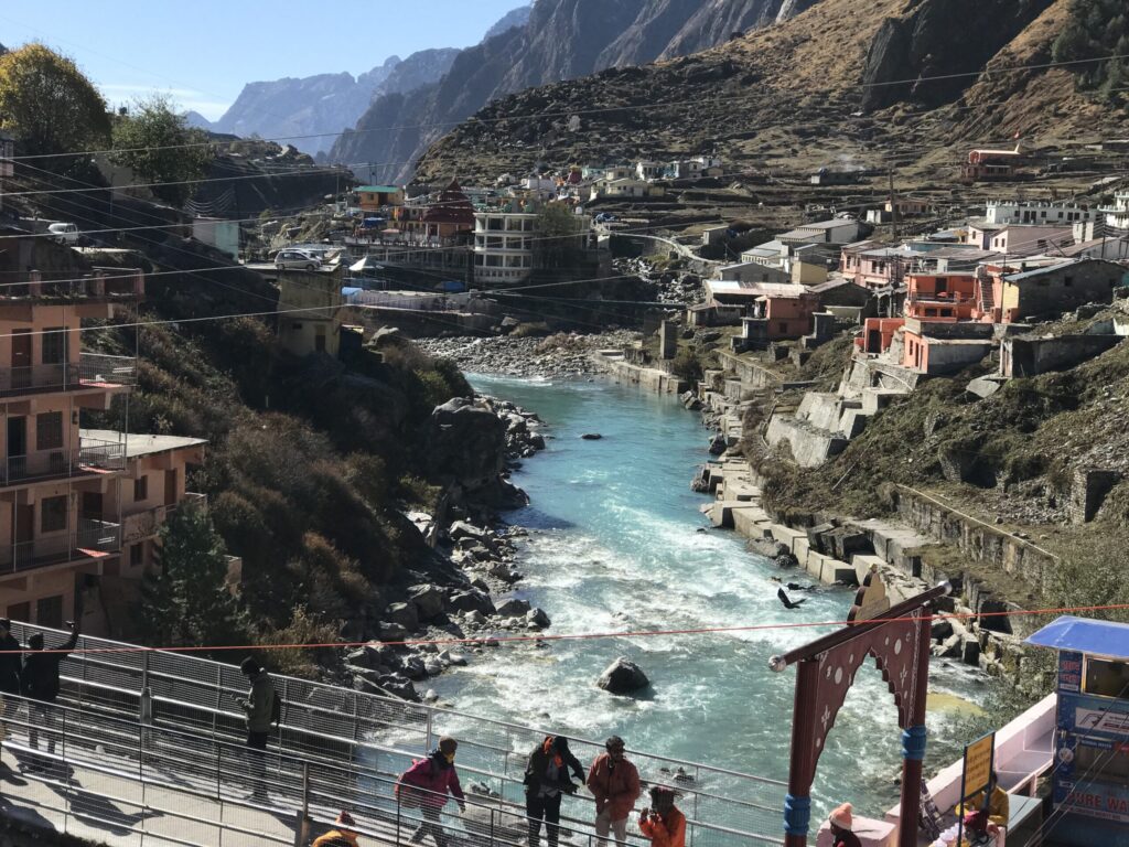Alaknanda River flowing through the town of Badrinath, with pilgrims crossing a bridge and mountain houses lining the riverbanks amid the Himalayan landscape.