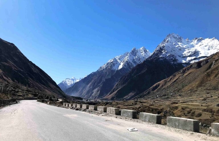 Wide mountain road leading through a Himalayan valley near Badrinath, with snow-covered peaks towering on both sides under a clear blue sky.