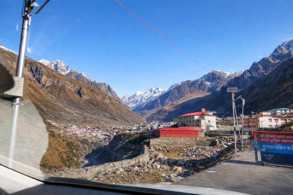 View of Badrinath town and surrounding snow-capped Himalayan peaks seen from inside a vehicle, with roads, buildings, and mountain slopes under a clear blue sky.