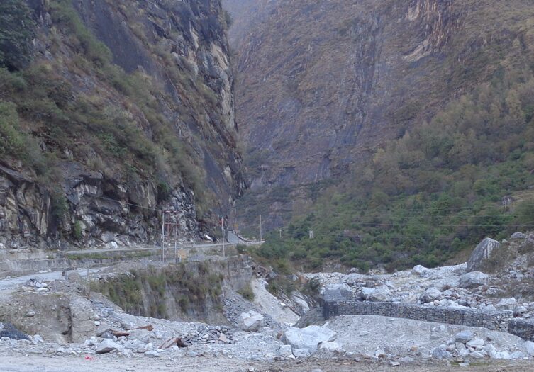 Mountain road carved along a steep rocky cliff in the Himalayas, with a dry riverbed, scattered boulders, and sparse greenery in a deep valley below.