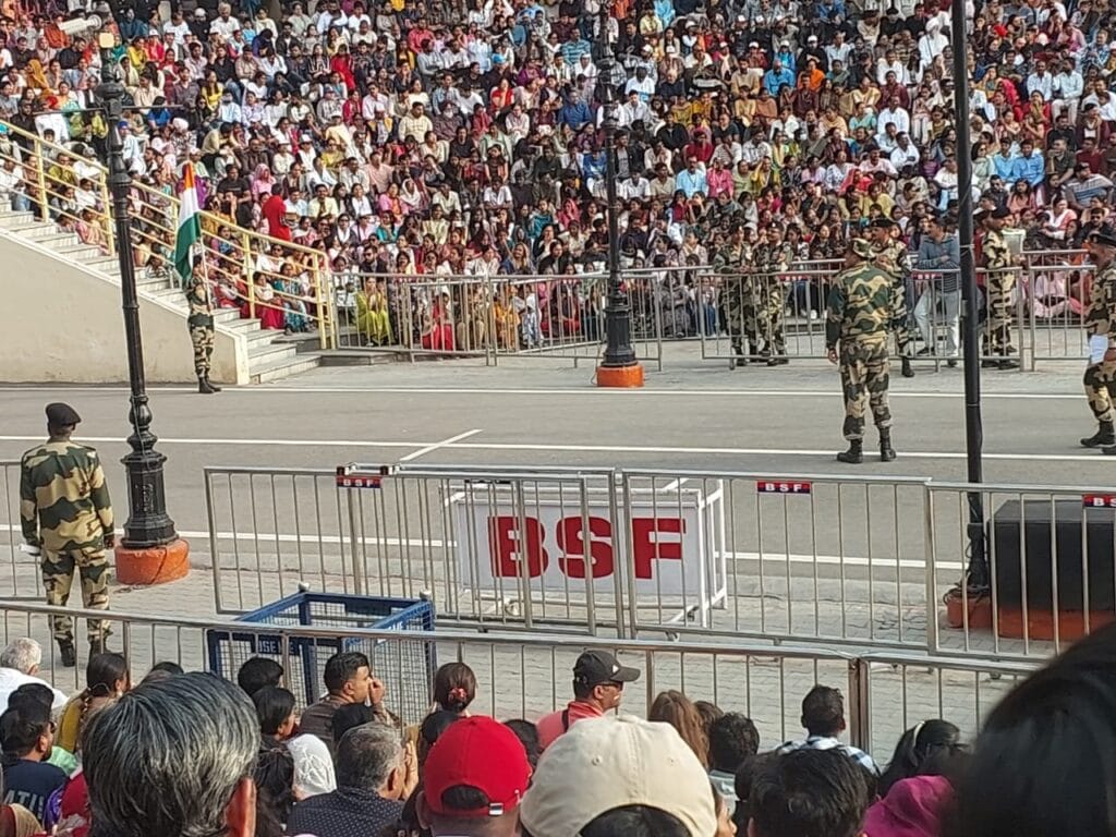 BSF soldiers standing guard near the parade area at Wagah Border, with a packed stadium of spectators watching the evening retreat ceremony from the stands.