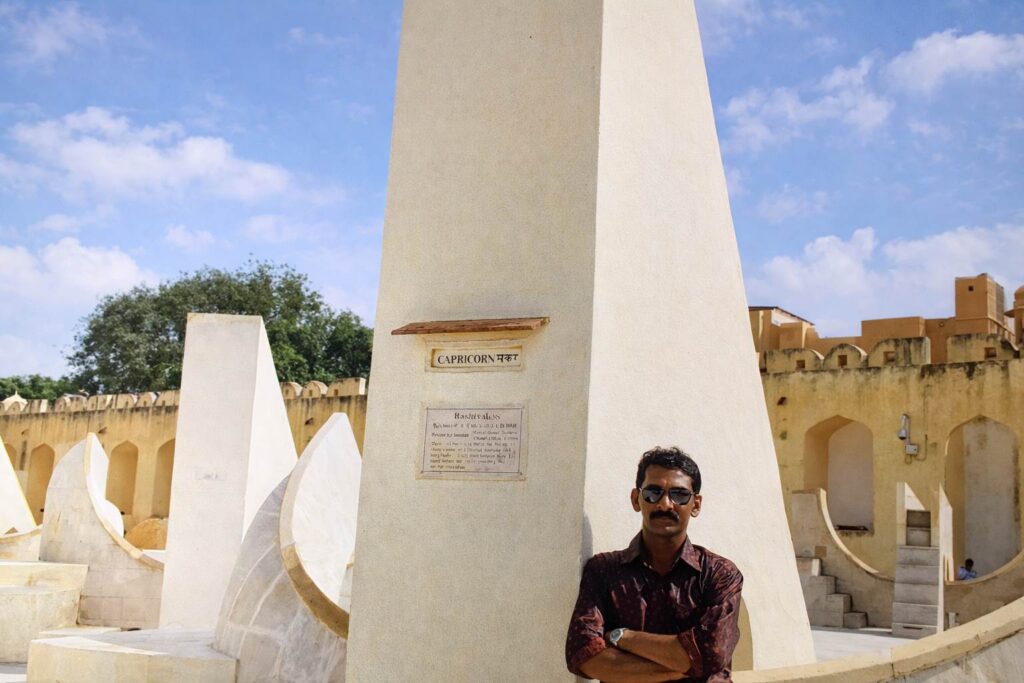 Man standing beside Capricorn instrument at Jantar Mantar Jaipur, symbolic alignment of zodiac sign with astronomical structure under blue sky