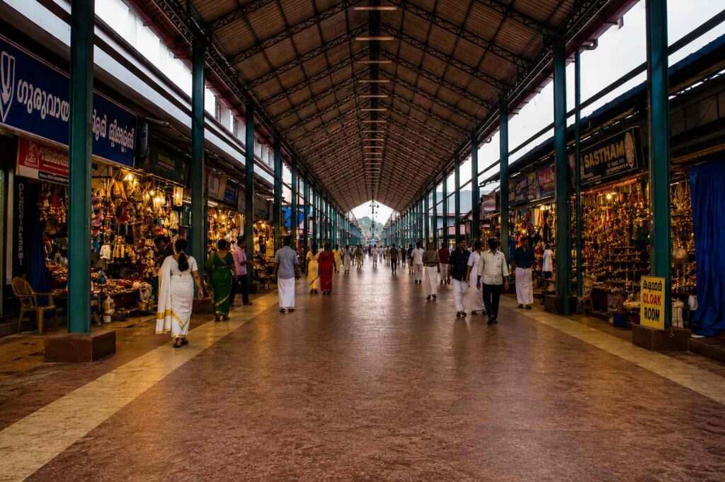 Covered shopping street near Guruvayoor Temple lined with traditional shops, brass lamps, devotional items, and people walking through the long corridor.