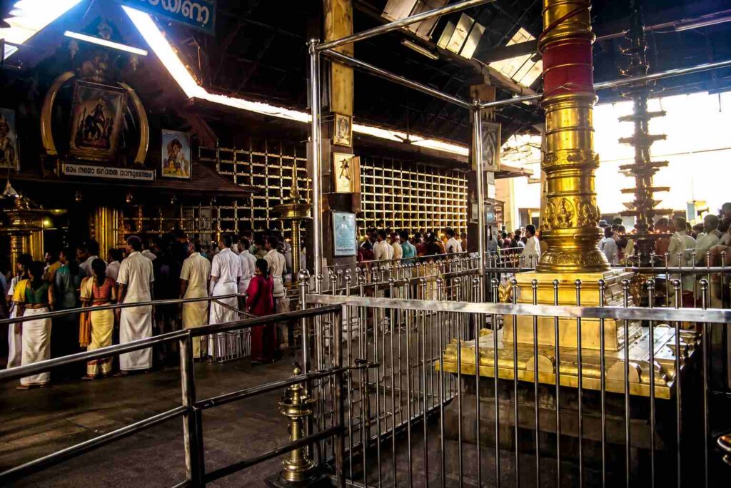 Devotees standing in queue near the golden deepasthambam inside Guruvayur Temple complex surrounded by metal railings and traditional Kerala-style architecture. 