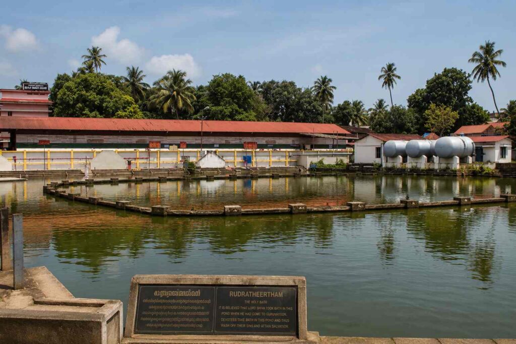 View of Rudratheertham pond near Guruvayoor Temple with calm green water, circular stone walkway, Kerala-style buildings, and coconut trees in the background.