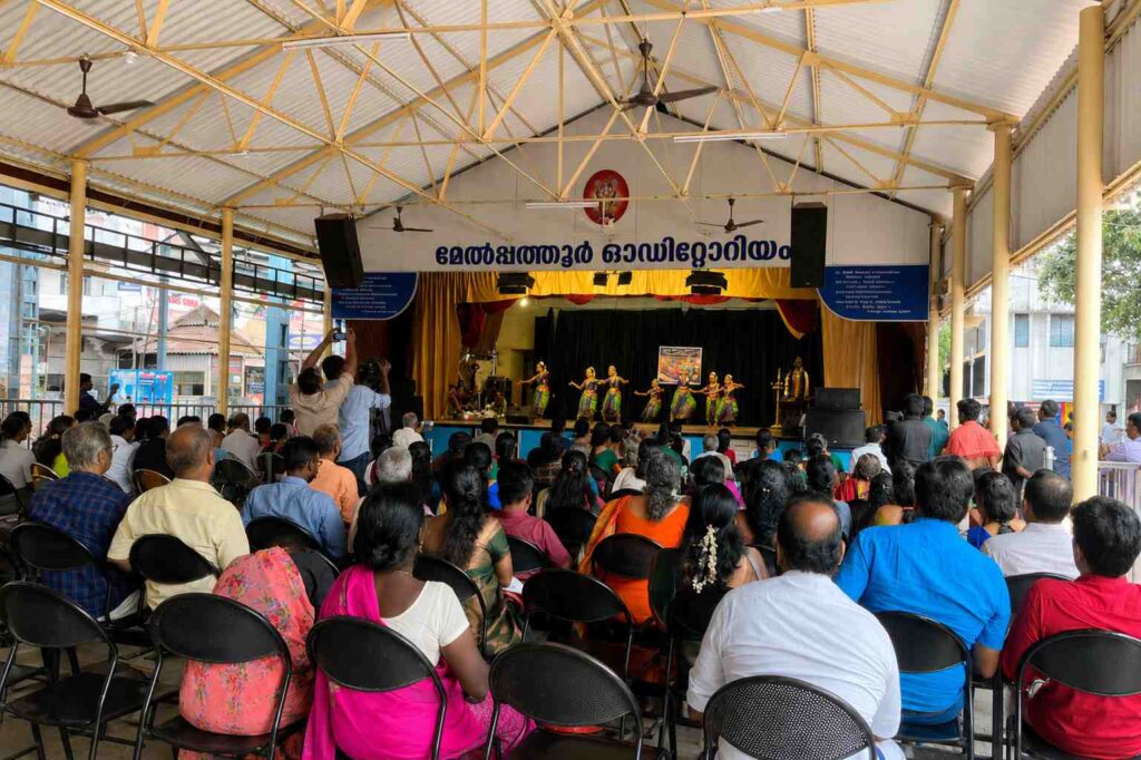 Audience watching a traditional cultural dance performance inside Melpathur Auditorium in Guruvayur, featuring a spacious covered hall and vibrant stage setting.
